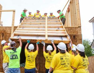 Ten volunteers pushing the frame of a new wall to the second floor where 6 volunteers are pulling the wall frame onto the second floor.