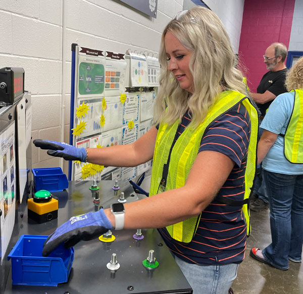 Female educator at the Whirlpool Manufacturing bootcamp testing equipment at manufacturing plant. She has long blonde hair, is smiling, and wearing a striped shirt with a safety vest over it, and protective gloves. Additional teachers are in the background.